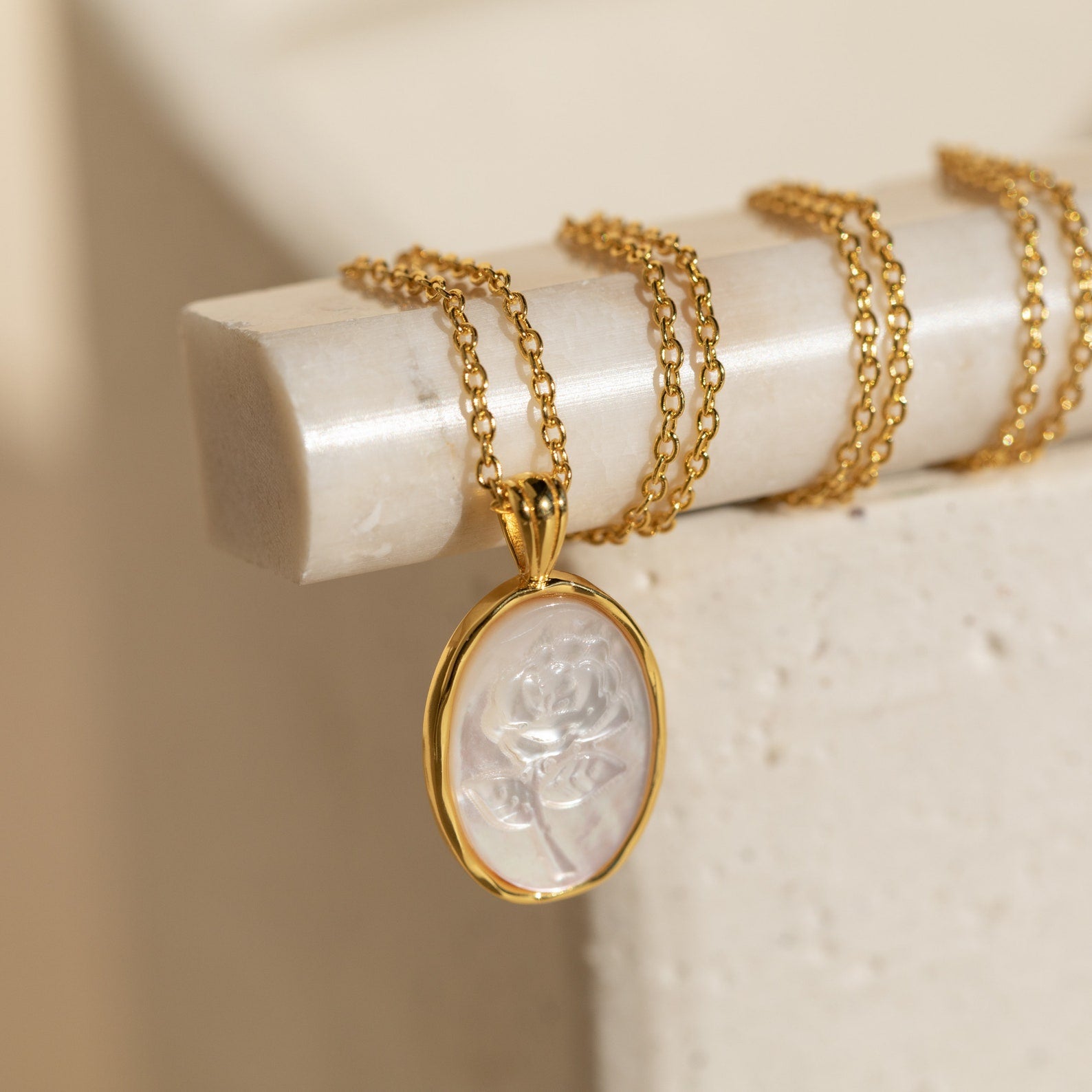 The Vintage Rose Necklace features a gold chain and an oval translucent pendant engraved with a rose, displayed on a cream surface—a lovely flower necklace perfect for Mother’s Day.