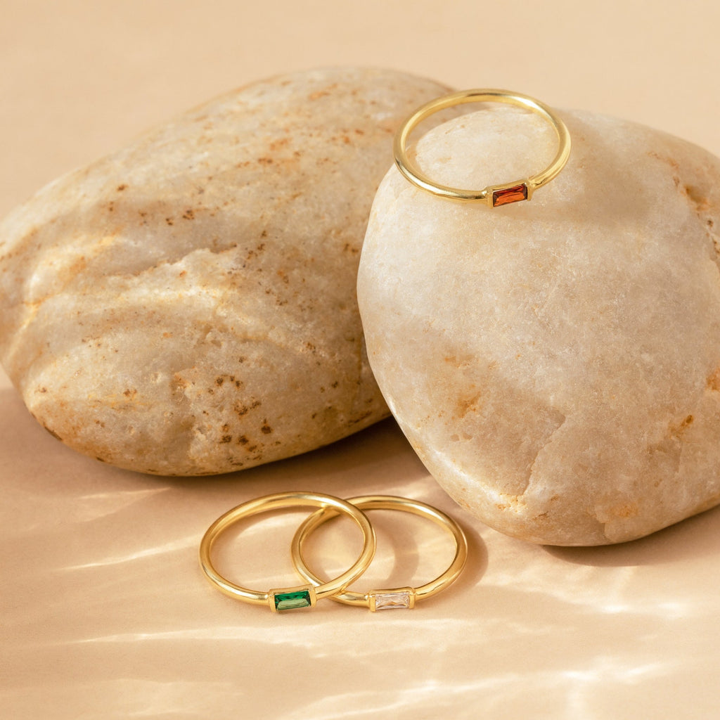 The Baguette Birthstone Ring, a gold stacking ring with colored gemstones, is displayed on and in front of smooth beige stones, showcasing the elegance of personalized jewelry.