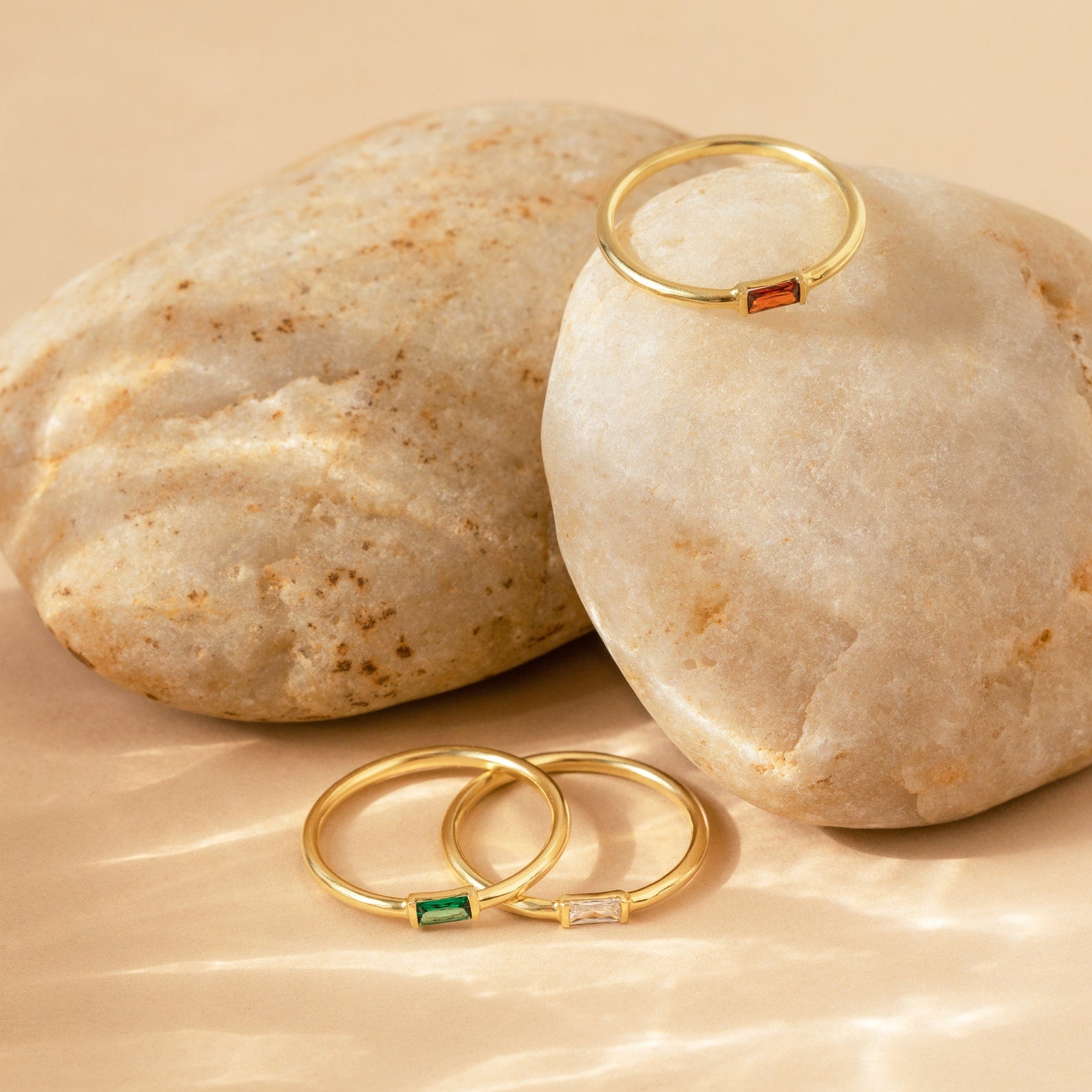 The Baguette Birthstone Ring, a gold stacking ring with colored gemstones, is displayed on and in front of smooth beige stones, showcasing the elegance of personalized jewelry.