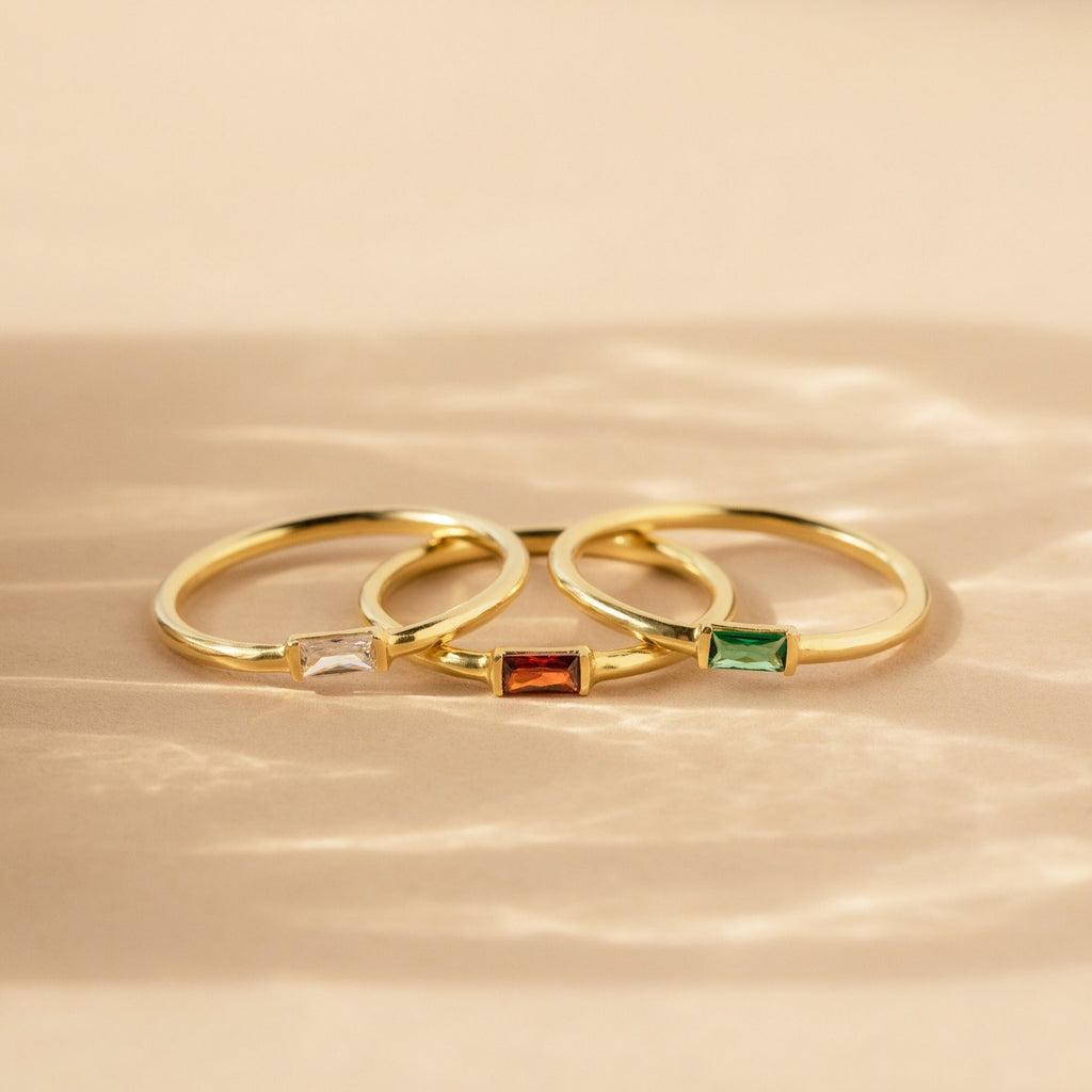 Three gold Baguette Birthstone Rings with rectangular white, red, and green stones rest on a beige surface in soft light. Personalize your style with this customizable ring set.