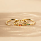 Three gold Baguette Birthstone Rings with rectangular white, red, and green stones rest on a beige surface in soft light. Personalize your style with this customizable ring set.