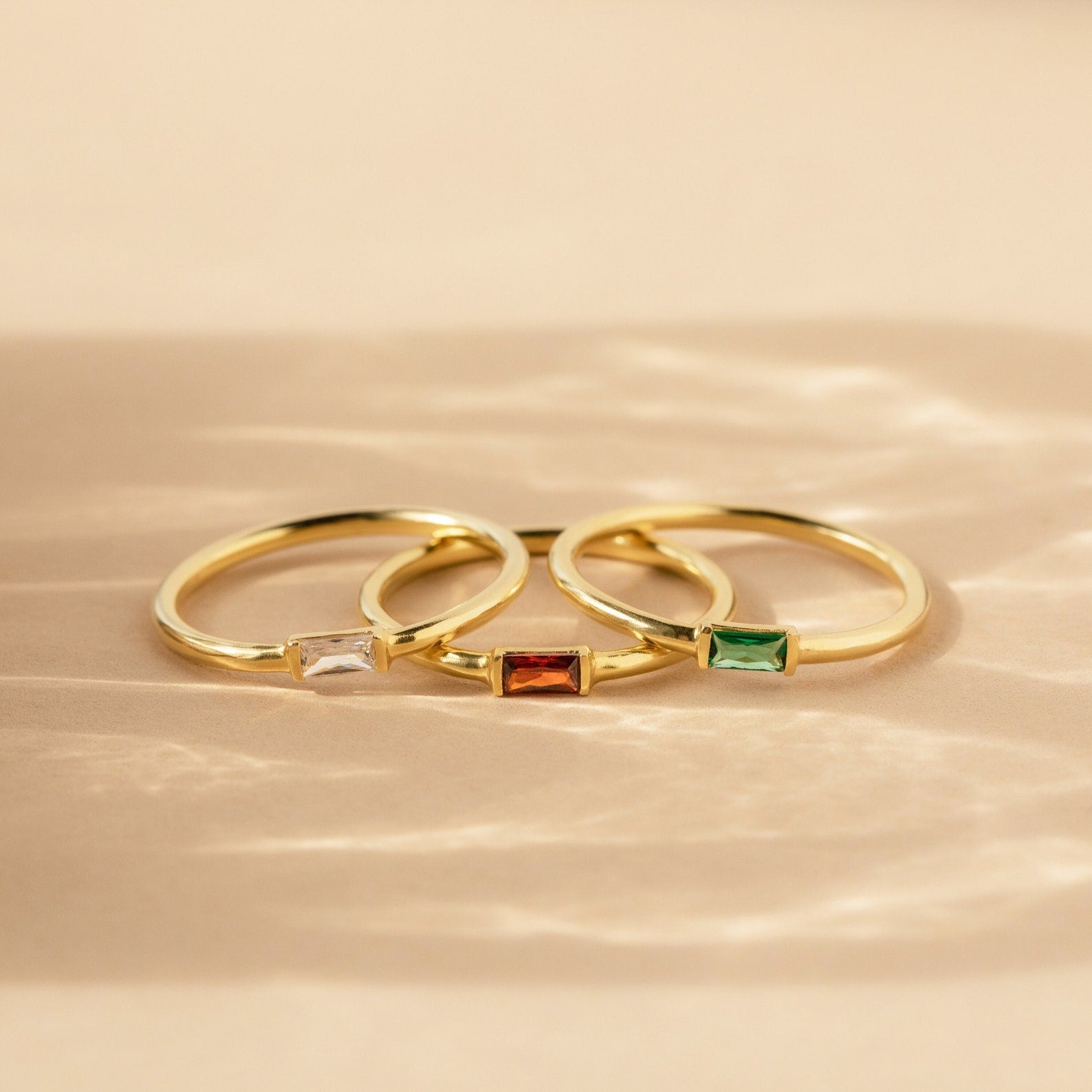 Three gold Baguette Birthstone Rings with rectangular white, red, and green stones rest on a beige surface in soft light. Personalize your style with this customizable ring set.