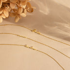 The Venice Tiny Name Bracelet, a delicate gold chain with a cursive name pendant, is beautifully displayed on a beige surface near dried flowers—perfect for layering with minimal jewelry.