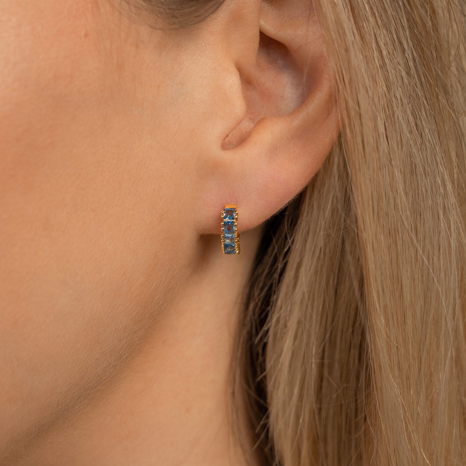 Close-up of a woman's ear wearing Genevieve Birthstone Huggies with blue gemstones—small gold earrings that make a beautiful gift for mom.