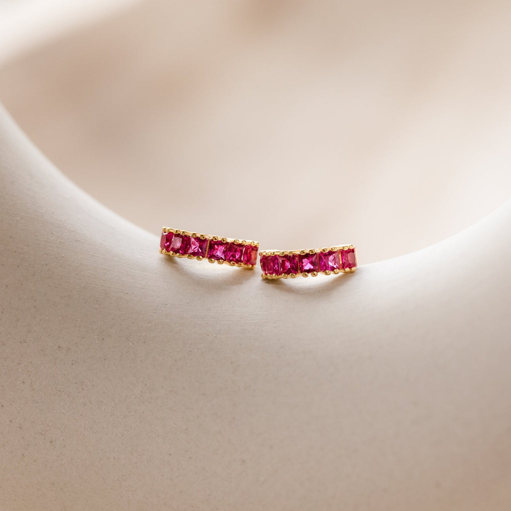 The Genevieve Birthstone Huggies in Ruby (July) are shown on a light, smooth surface—making them a charming gift for mom.