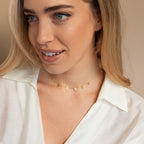 A woman smiles and looks to the side, wearing a white blouse paired with the Apollo Letter Name Necklace spelling "EVERY" in gold.