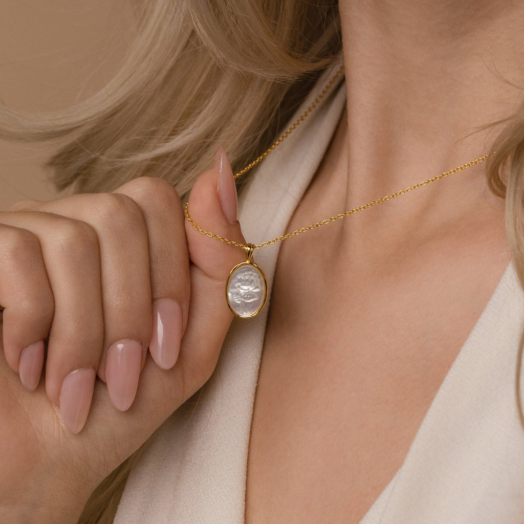 A woman with nude nails wears a cream top and holds the Vintage Rose Necklace featuring an oval pendant—a graceful floral piece, perfect for Mother’s Day.