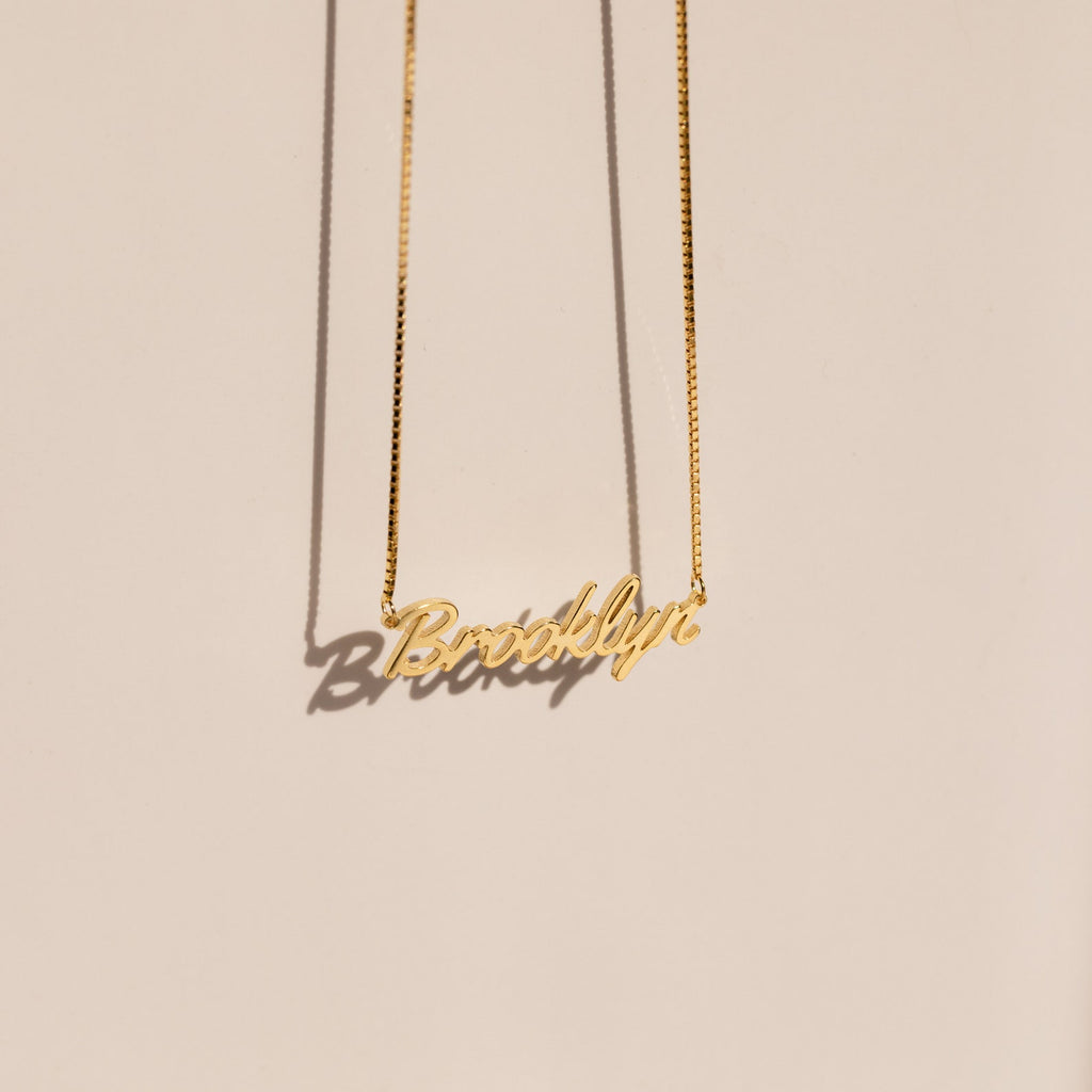 The Casella Name Necklace in Box Chain features a personalized pendant in a dainty script font, casting a shadow on a light beige background—an elegant custom gift for any occasion.