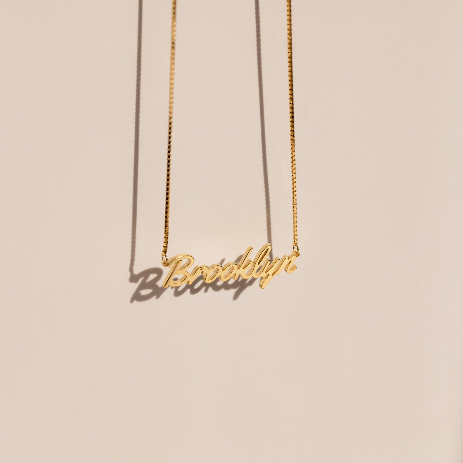 The Casella Name Necklace in Box Chain features a personalized pendant in a dainty script font, casting a shadow on a light beige background—an elegant custom gift for any occasion.