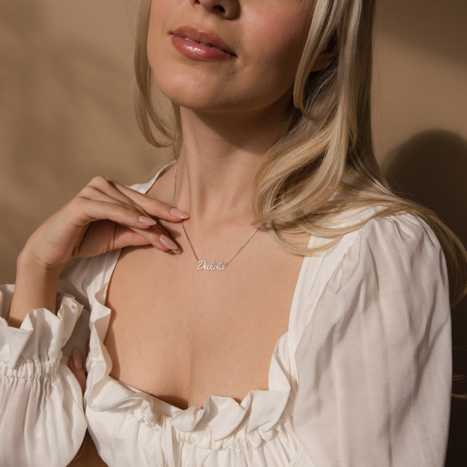 A woman in a white blouse touches a dainty script font pendant, elegantly showcasing the Casella Name Necklace in Box Chain against a beige background.