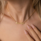 The Casella Name Necklace in Box Chain features a dainty script spelling Brooklyn, worn by a woman with light skin and manicured nails—an ideal personalized gift for any occasion.