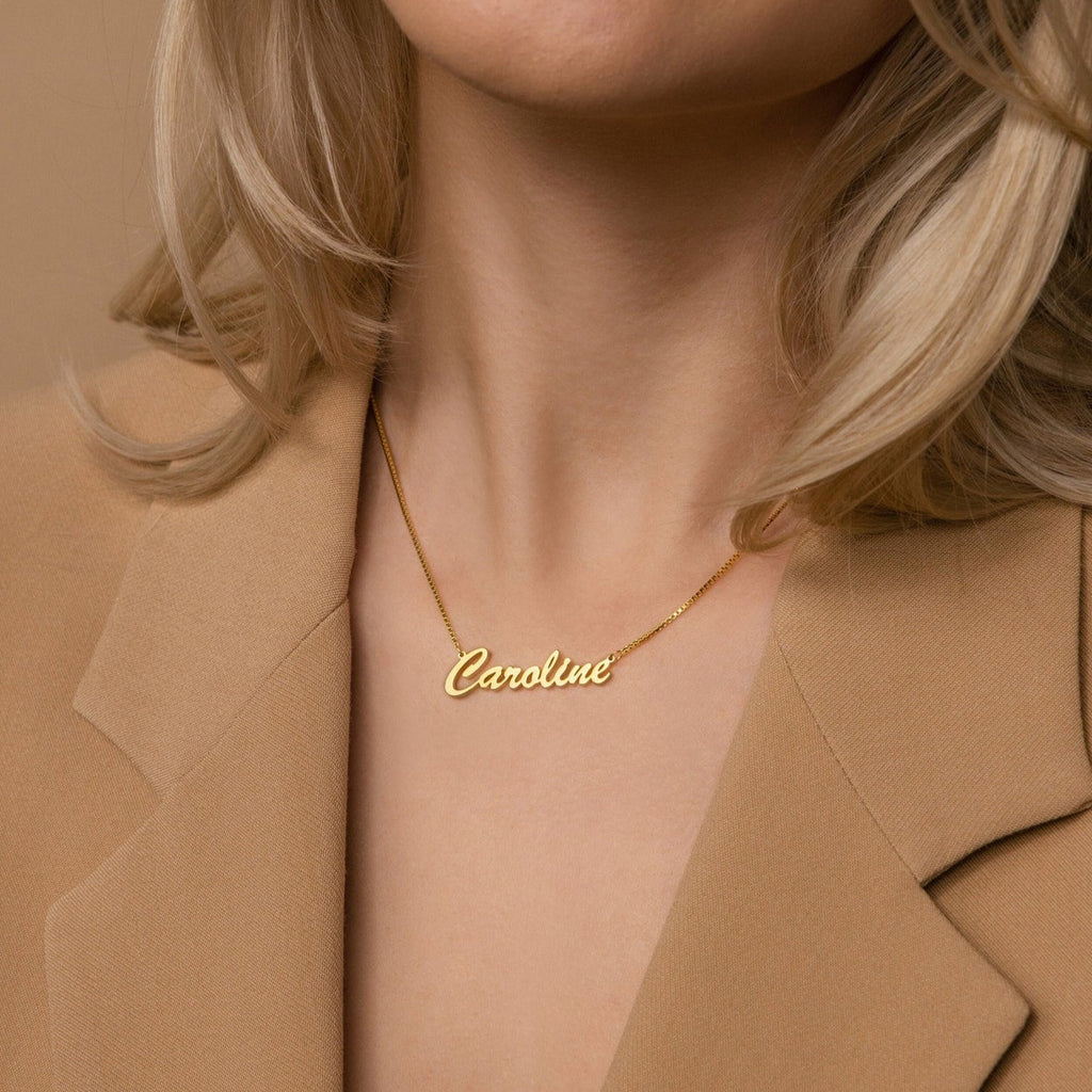 A woman wears the Natural Name Necklace in Box Chain customized with "Caroline," paired with a beige blazer. This elegant, personalized necklace makes a thoughtful gift.
