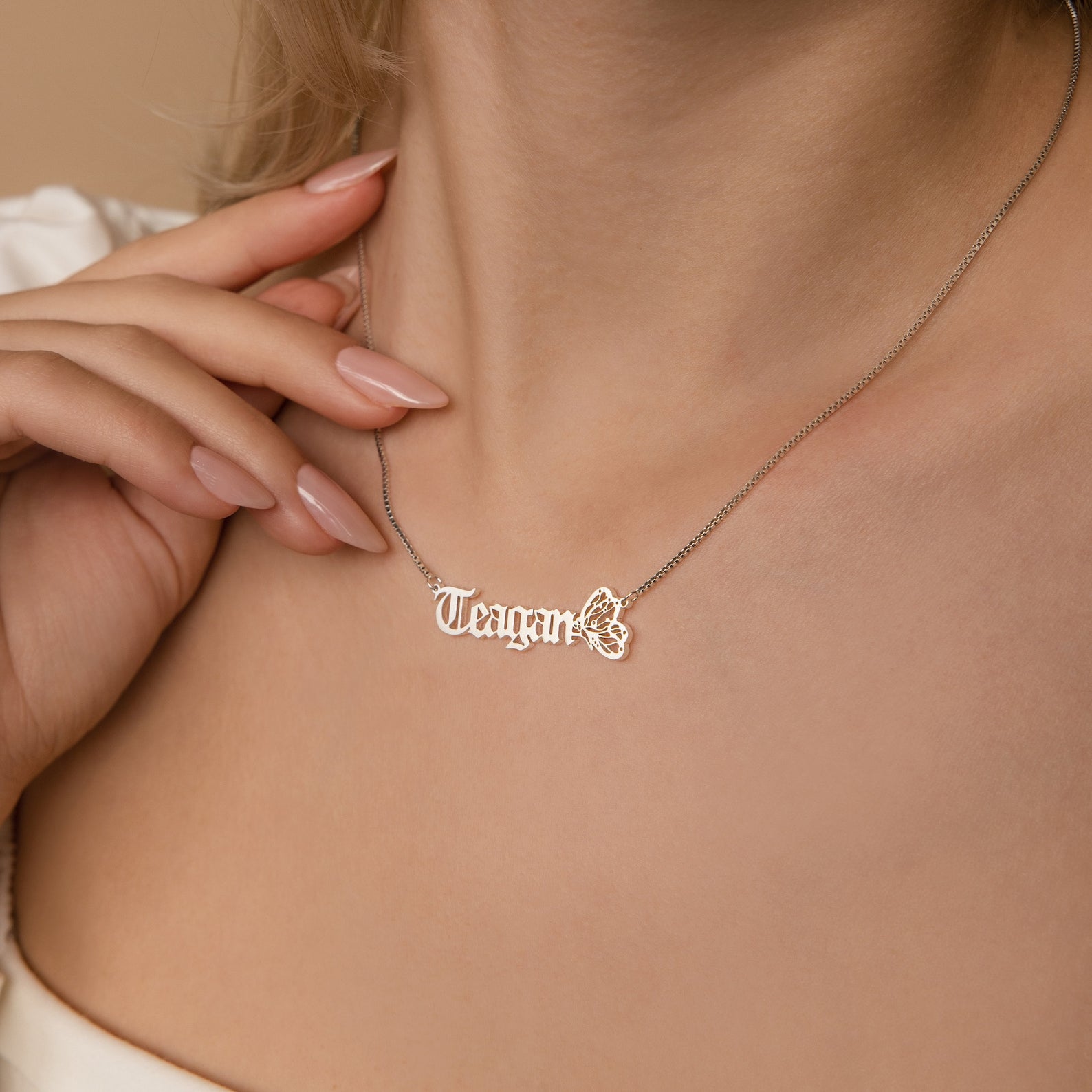 A woman wears the Butterfly Name Necklace in Box Chain personalized with the name Teagan and a butterfly charm, gently holding it near her collarbone.