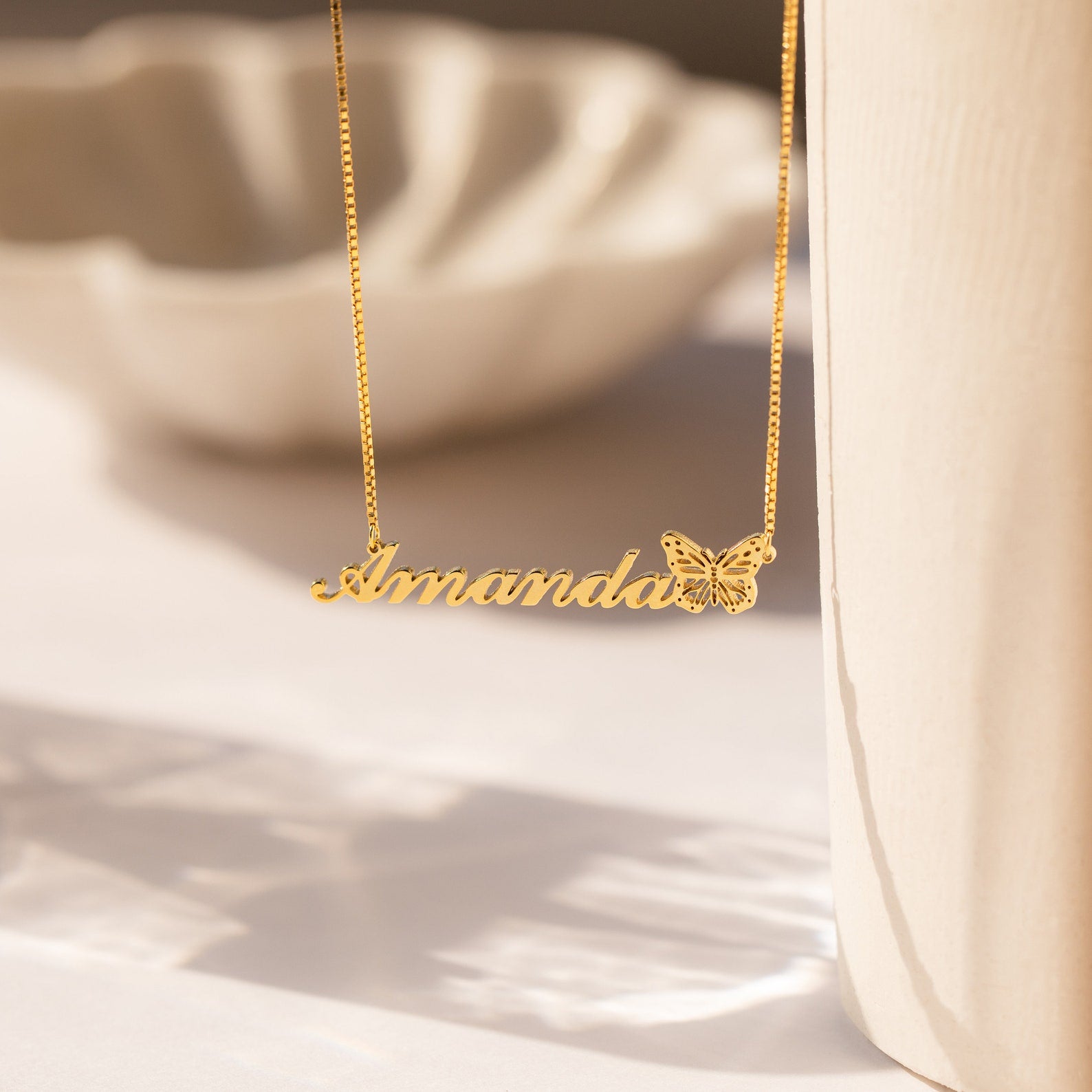 The Butterfly Name Necklace in Box Chain features the name Amanda and a delicate butterfly charm, beautifully catching soft sunlight.