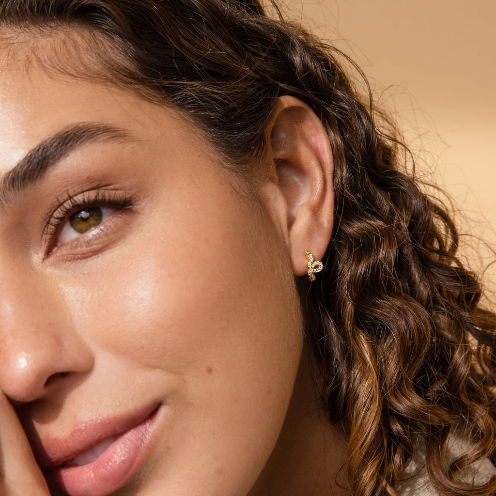 A woman with curly hair smiles, resting her face on her hand, showcasing trendy jewelry featuring the Cobra Snake Huggies gold earrings.