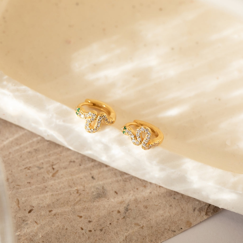 Cobra Snake Huggies—gold hoop earrings set with green and clear gemstones reminiscent of emeralds and diamonds—rest on a cream, textured surface in soft light, making them an ideal pick for trend-forward jewelry lovers.