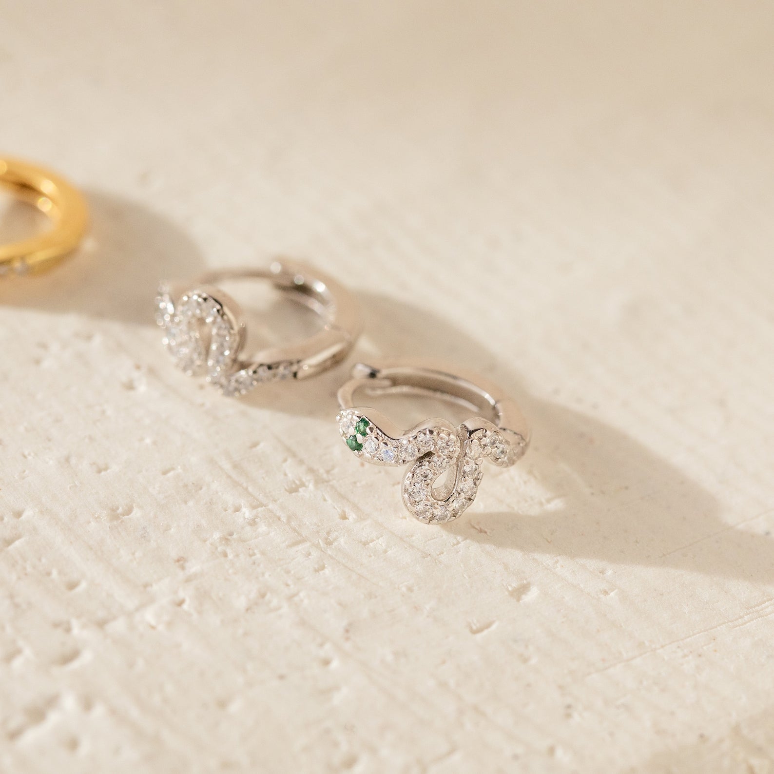 The Cobra Snake Huggies feature a trendy silver snake design with a sparkling green stone as the serpent's eye, perfect for stylish jewelry lovers.