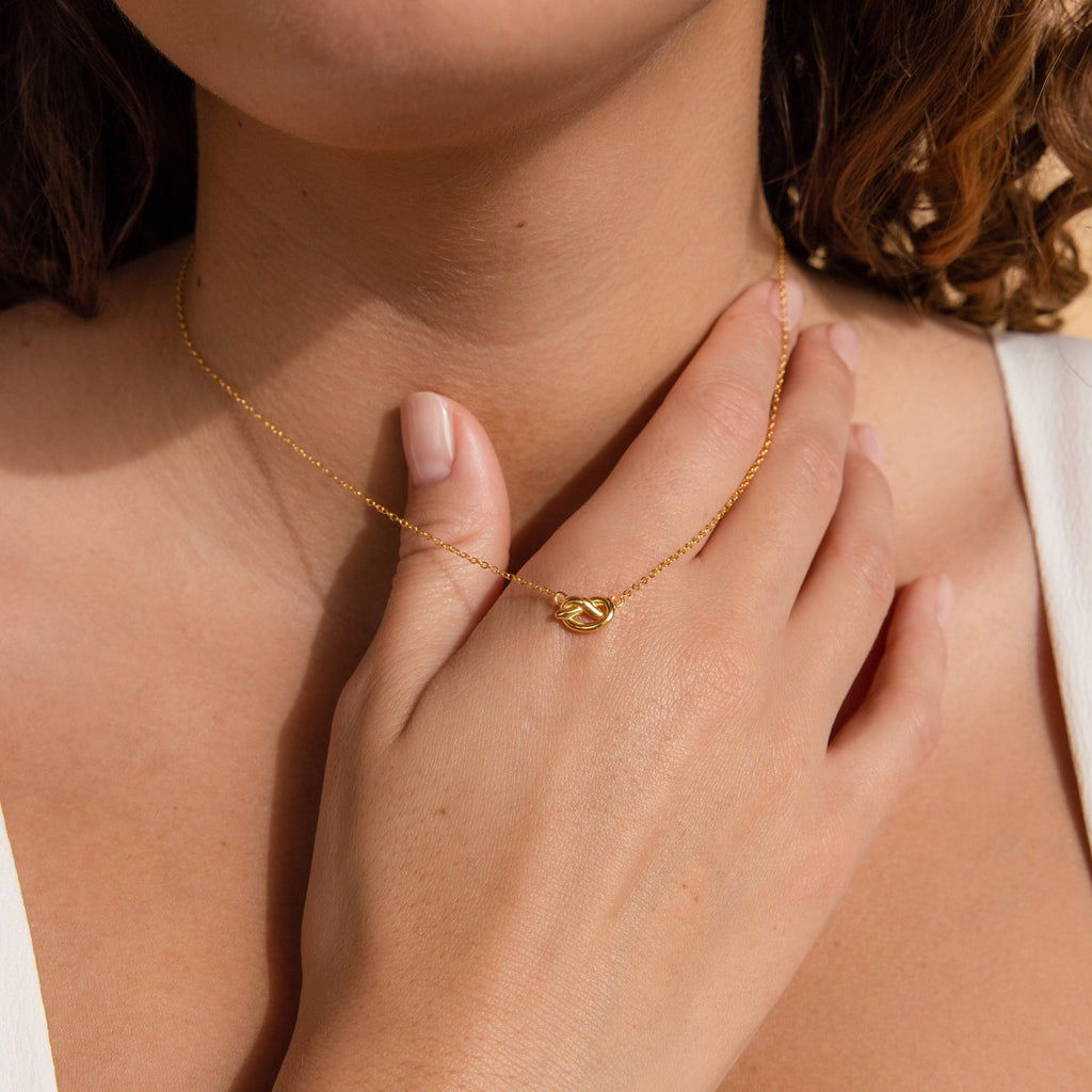 A woman touches her chest, wearing the Love Knot Necklace—a gold chain with a knot pendant that symbolizes unity and makes a meaningful wedding gift.