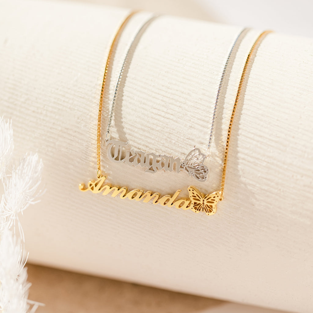 The Butterfly Name Necklace in Box Chain is shown in gold and silver variants with butterfly charms on a cream surface—ideal as a personalized necklace or unique name pendant.