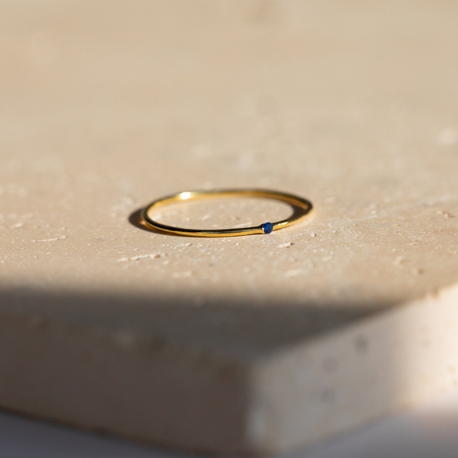 The Thin Mini Birthstone Ring, featuring a delicate blue stone, sits on a beige textured surface—an elegant piece of minimalist jewelry and an ideal Christmas gift.