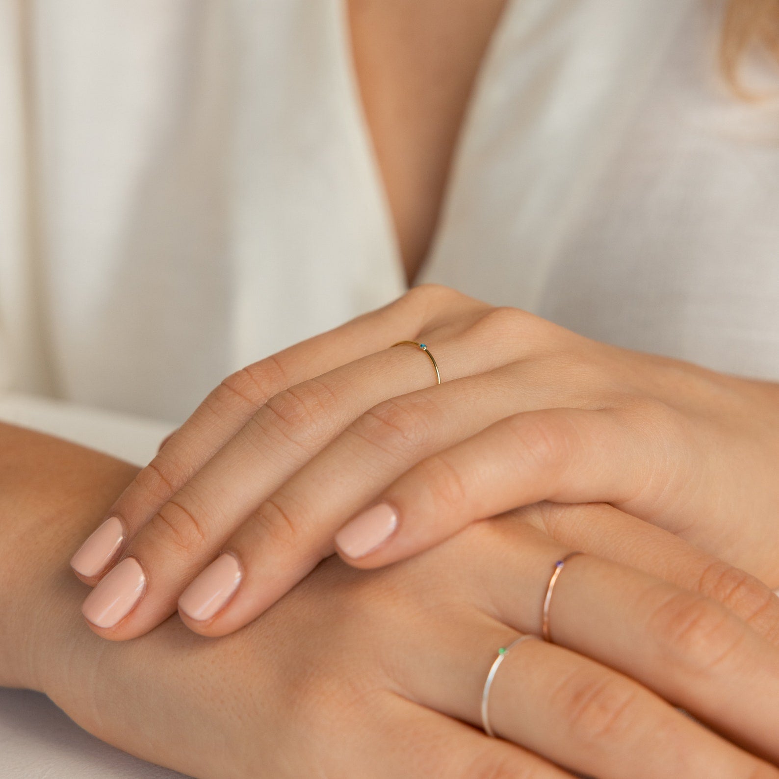 Close-up of hands with light pink manicured nails wearing minimalist jewelry, featuring the Thin Mini Birthstone Ring—an ideal Christmas gift—resting on a white surface.