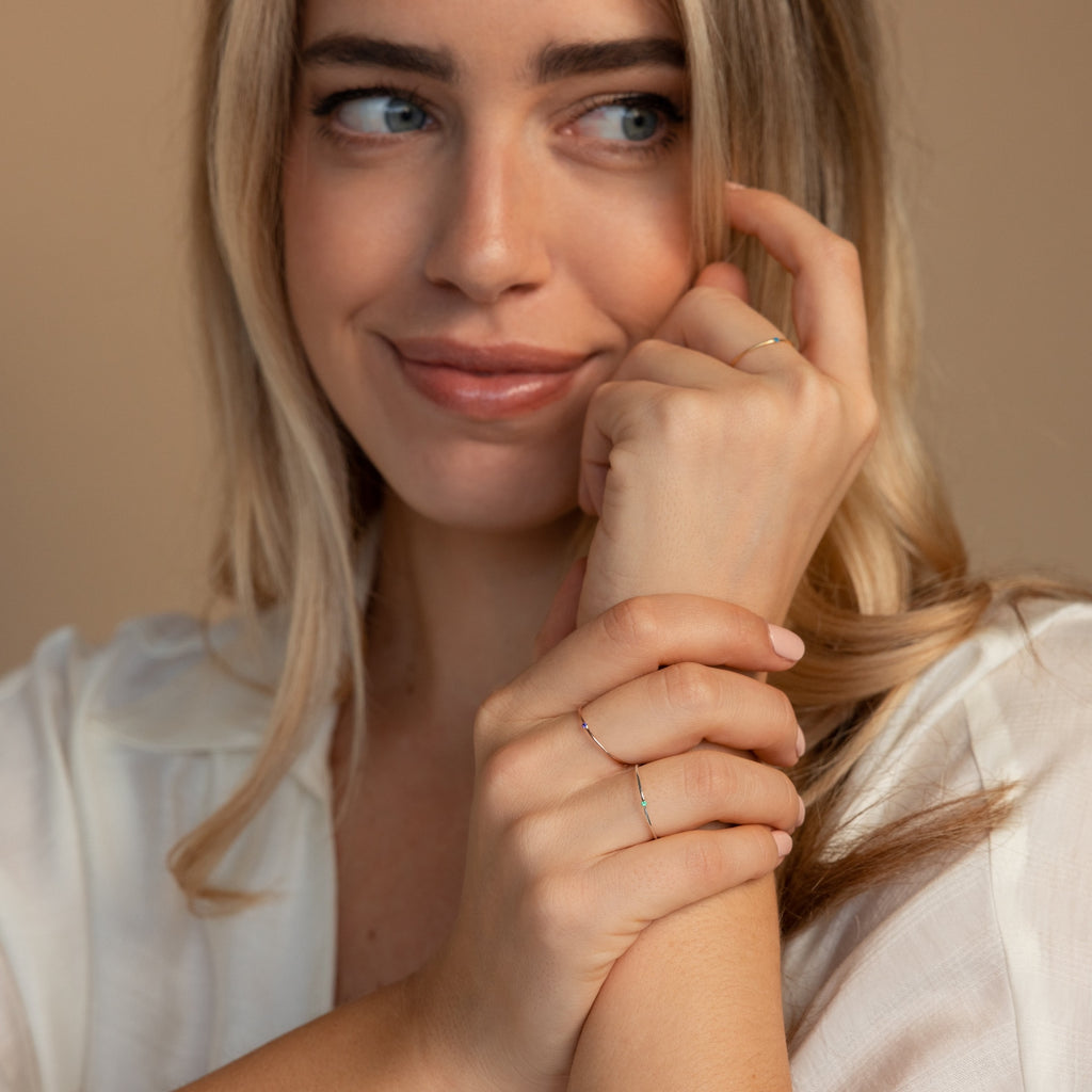 A woman with long blonde hair smiles gently, wearing a white blouse and the Thin Mini Birthstone Ring—an elegant, minimalist piece perfect as a Christmas gift.