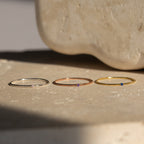 Three delicate Thin Mini Birthstone Rings in silver, rose gold, and gold with tiny colored gems rest on a textured surface—a minimalist jewelry piece and thoughtful Christmas gift.