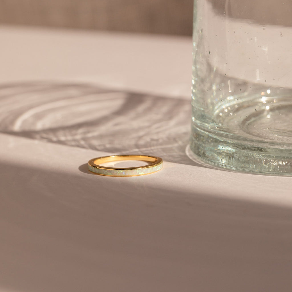 The White Opal Inlay Band rests on a white surface next to a clear glass, its elegant design highlighted by sunlight and subtle shadows.