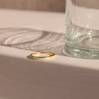 The White Opal Inlay Band rests on a white surface next to a clear glass, its elegant design highlighted by sunlight and subtle shadows.