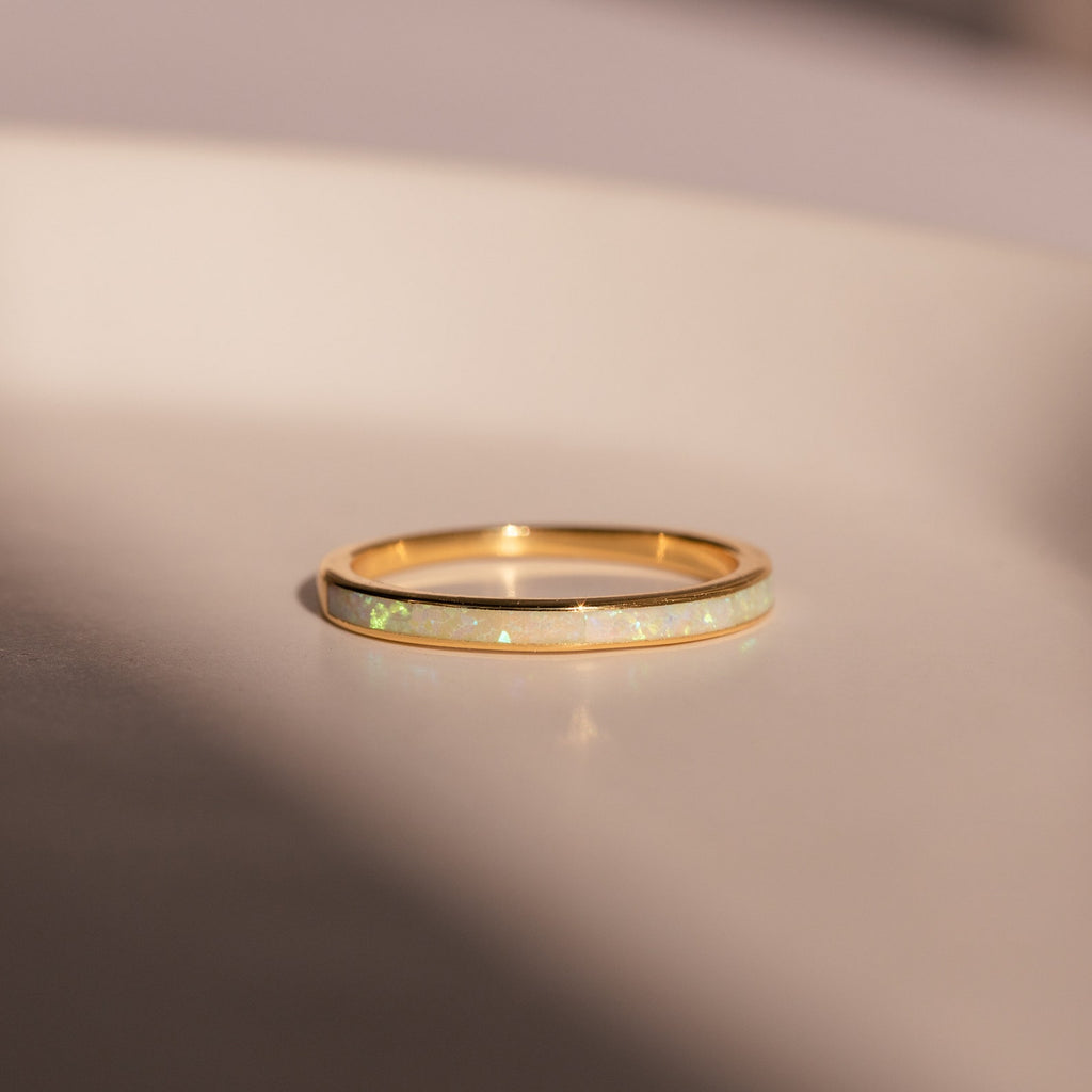 The White Opal Inlay Band, a thin gold ring featuring a white opal inlay, rests on a smooth, light-colored surface in soft lighting, highlighting the delicate beauty of this gemstone piece.