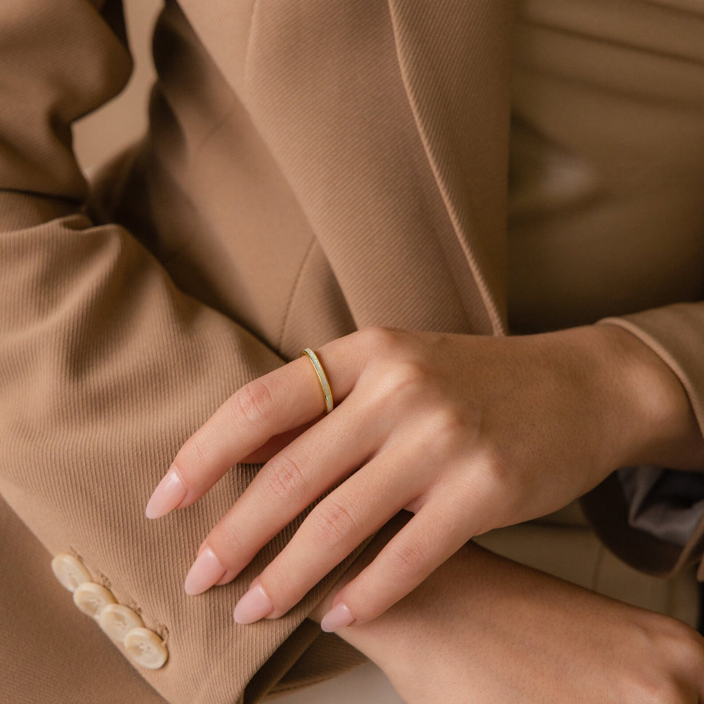 A hand with nude nail polish showcases the White Opal Inlay Band, resting on a tan suit sleeve with white buttons.