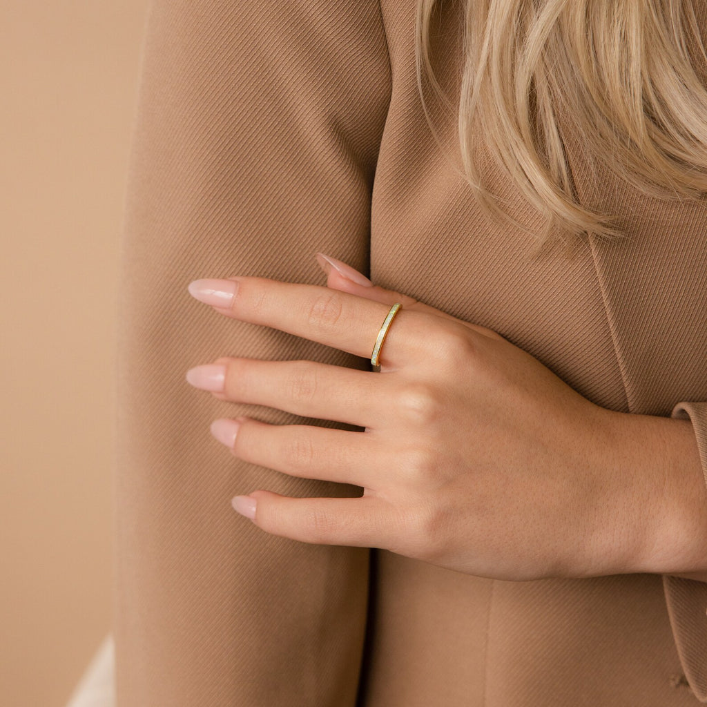 A hand with a nude manicure wearing the White Opal Inlay Band in 18K Gold rests on a tan, textured blazer sleeve.