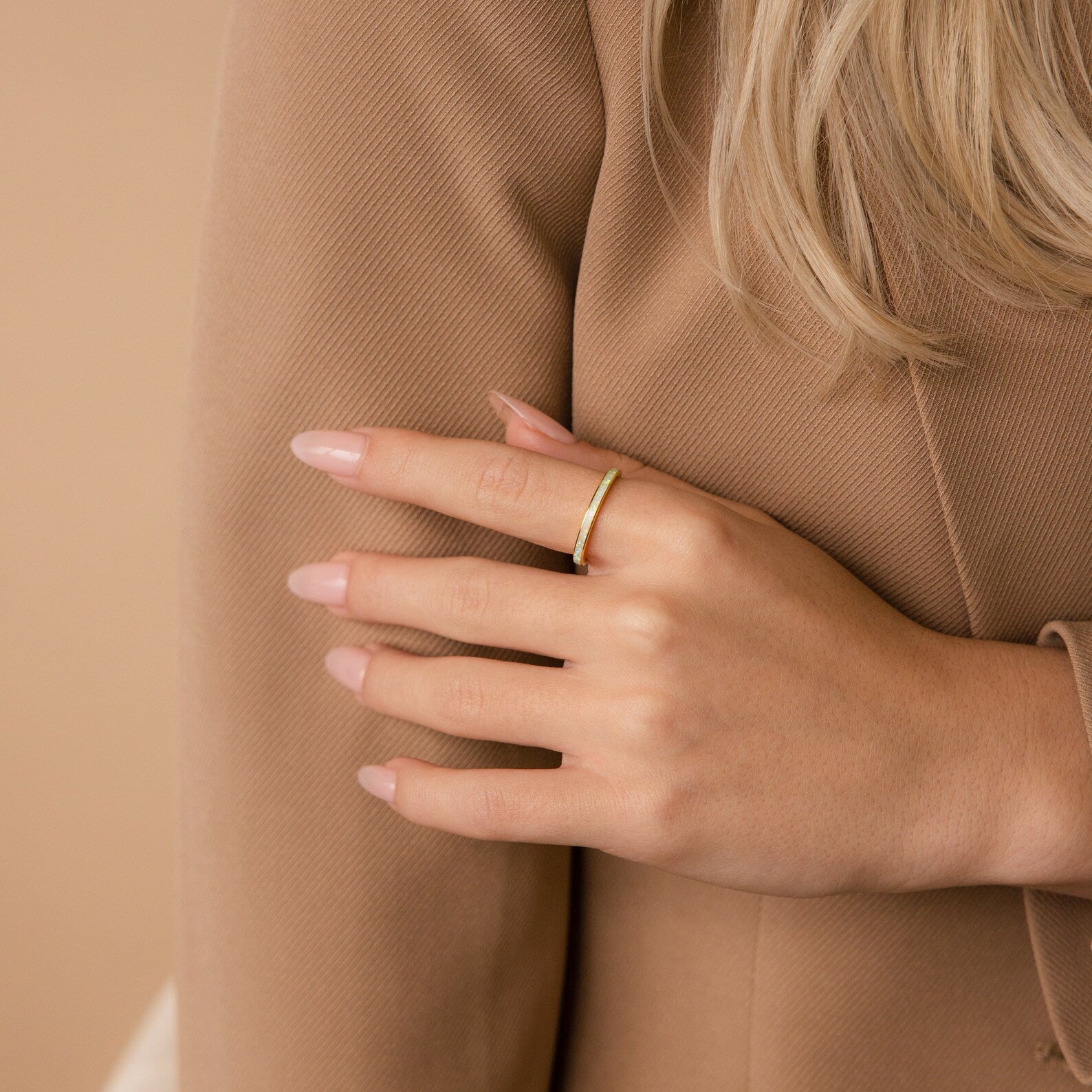 A hand with a nude manicure wearing the White Opal Inlay Band in 18K Gold rests on a tan, textured blazer sleeve.