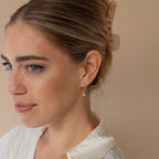 A woman with light hair in a claw clip and a white top looks to the side, showcasing elegant Isha Pearl Drop Earrings.