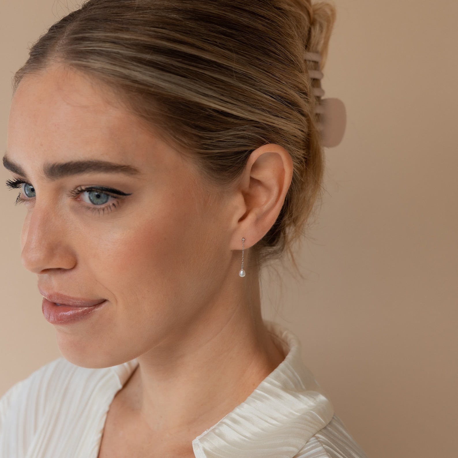 A woman with light hair in a claw clip and a white top looks to the side, showcasing elegant Isha Pearl Drop Earrings.