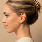 Woman with light hair in a bun, wearing Ericka Diamond Huggies with a subtle dangling charm, facing left against a beige background.