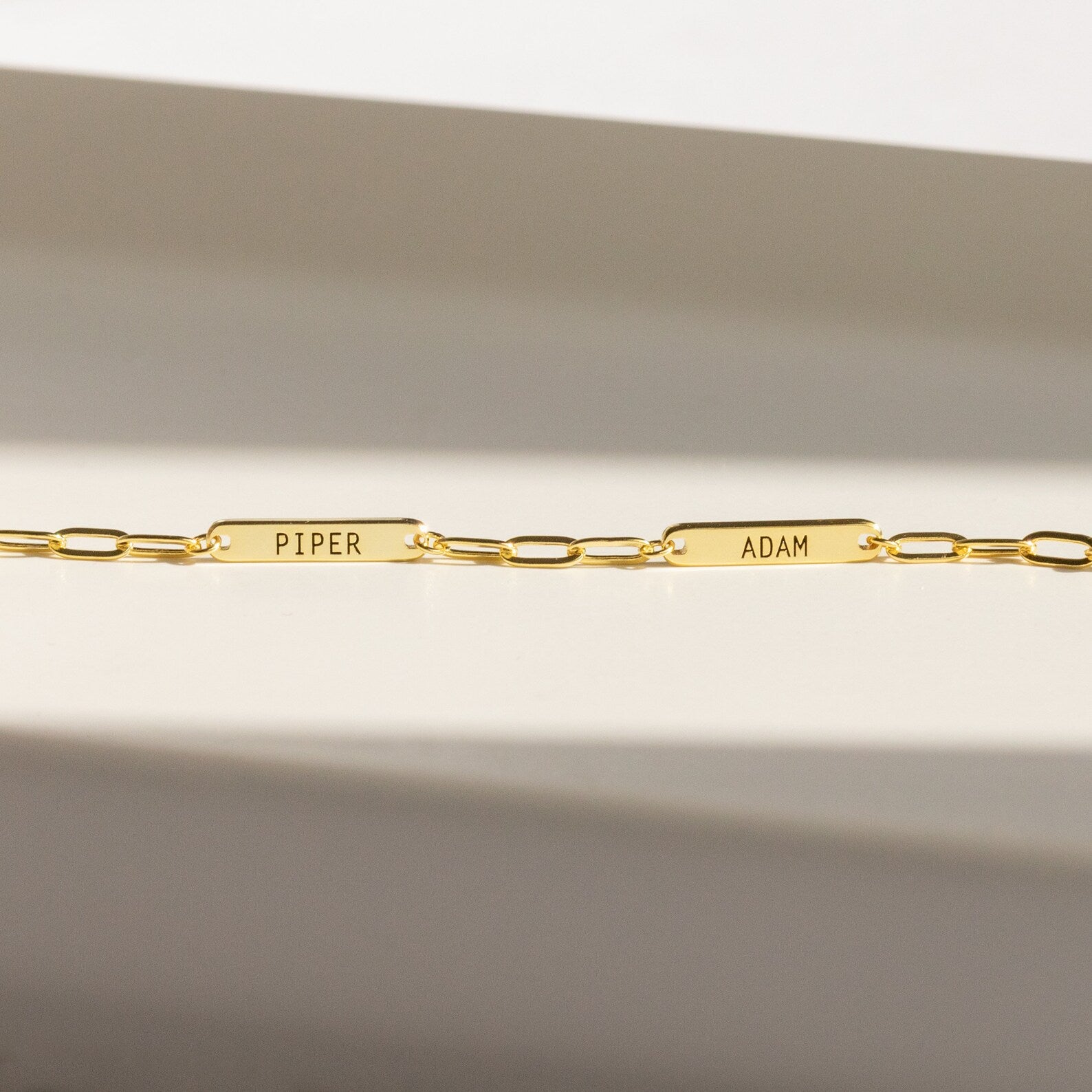 The Mini Name Bar Necklace features two rectangular name plates engraved with PIPER and ADAM, evoking the style of a personalized bar necklace, displayed against a neutral background.