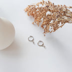 The Ericka Diamond Huggies in Sterling Silver are displayed on a white surface beside dried flowers and a round white object.