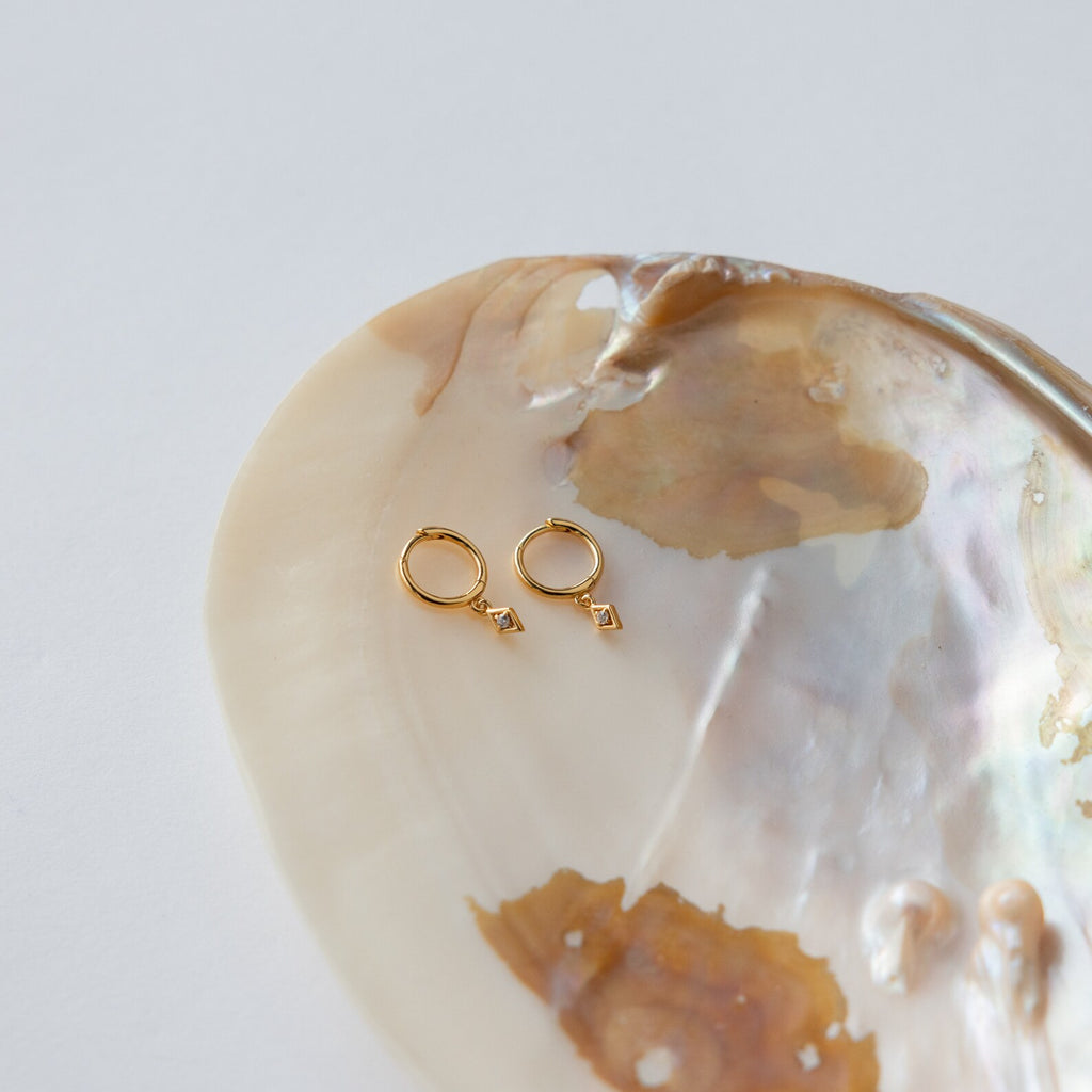 Ericka Diamond Huggies in 18K Gold with small geometric charms rest on an iridescent seashell against a white background.