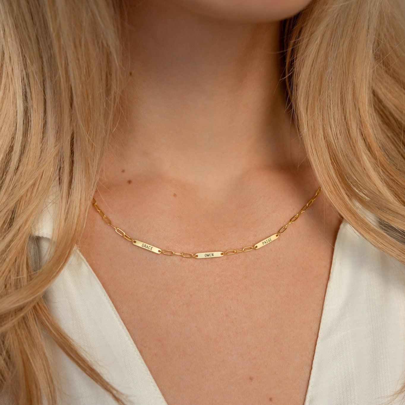 A woman wearing the Mini Name Bar Necklace, featuring three engraved rectangular charms, styled over a cream blouse.