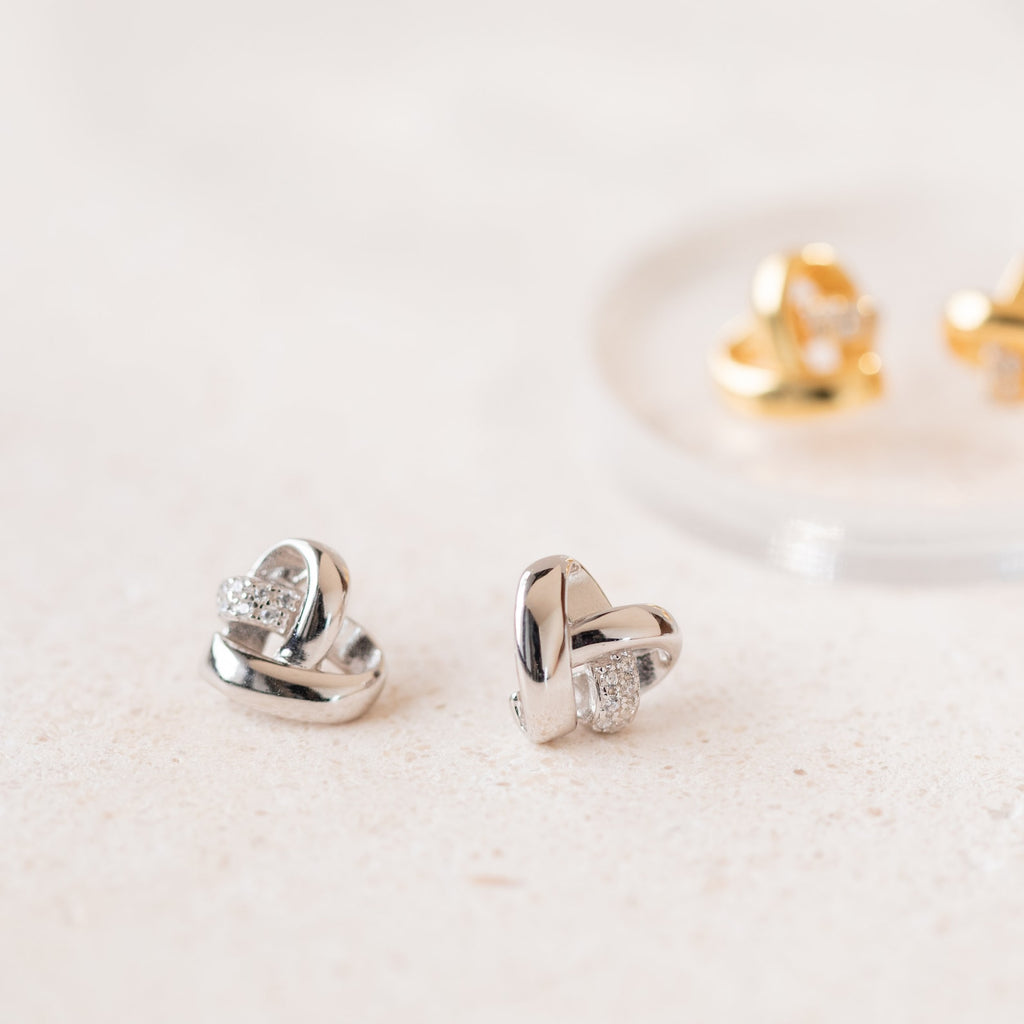 Two pairs of Heart Knot Studs—one silver with small crystals, one gold—feature a delicate knot design and are displayed on a light surface.