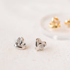 Two pairs of Heart Knot Studs—one silver with small crystals, one gold—feature a delicate knot design and are displayed on a light surface.