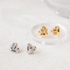 Heart Knot Studs in gold and silver, featuring a delicate knot design and small diamond charms, displayed on a light, textured surface.