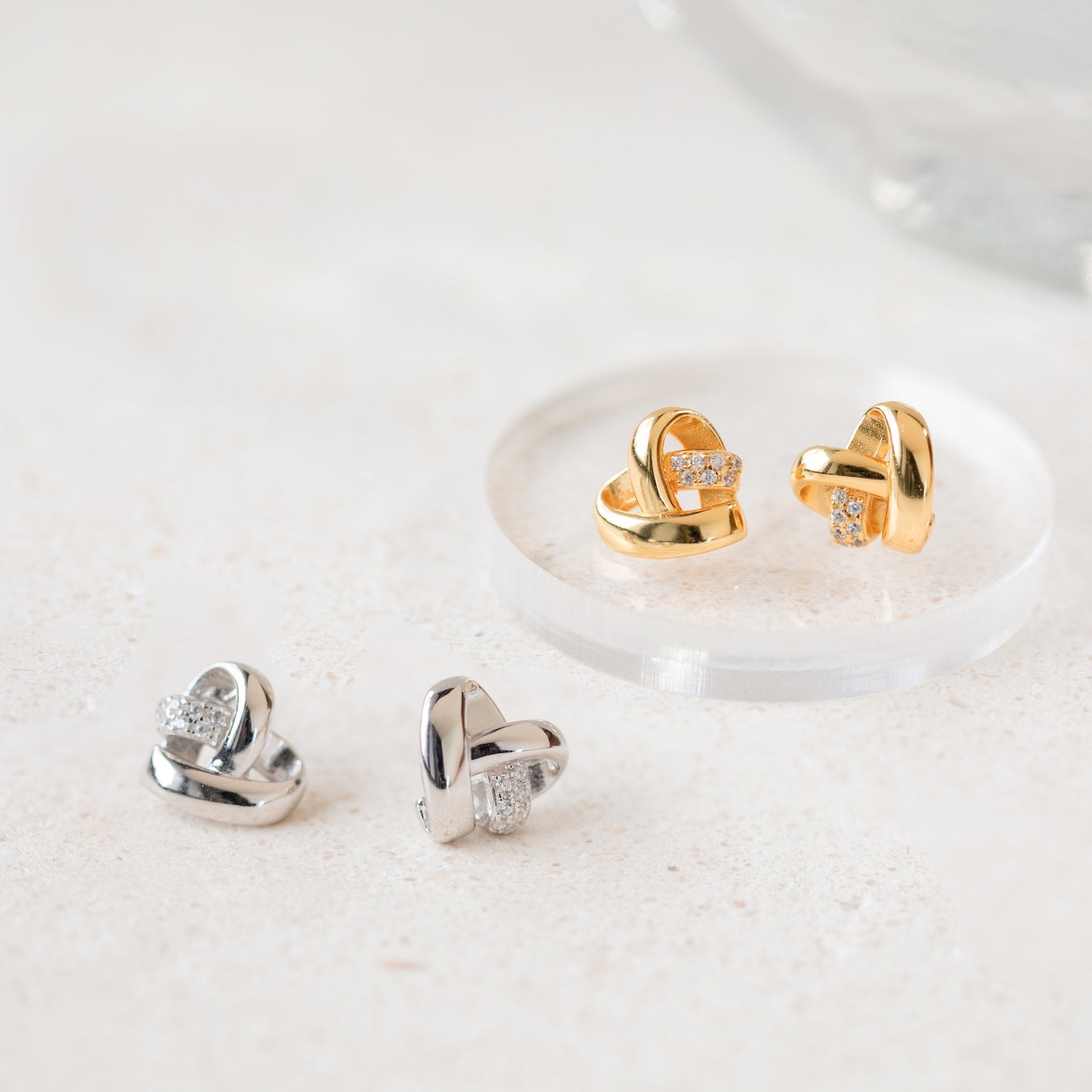 Heart Knot Studs in gold and silver, featuring a delicate knot design and small diamond charms, displayed on a light, textured surface.