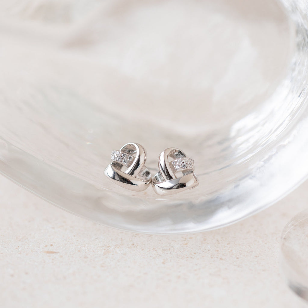 Heart Knot Studs: silver heart-shaped earrings with a knot design and small crystals, displayed on clear glass.
