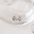 Heart Knot Studs: silver heart-shaped earrings with a knot design and small crystals, displayed on clear glass.