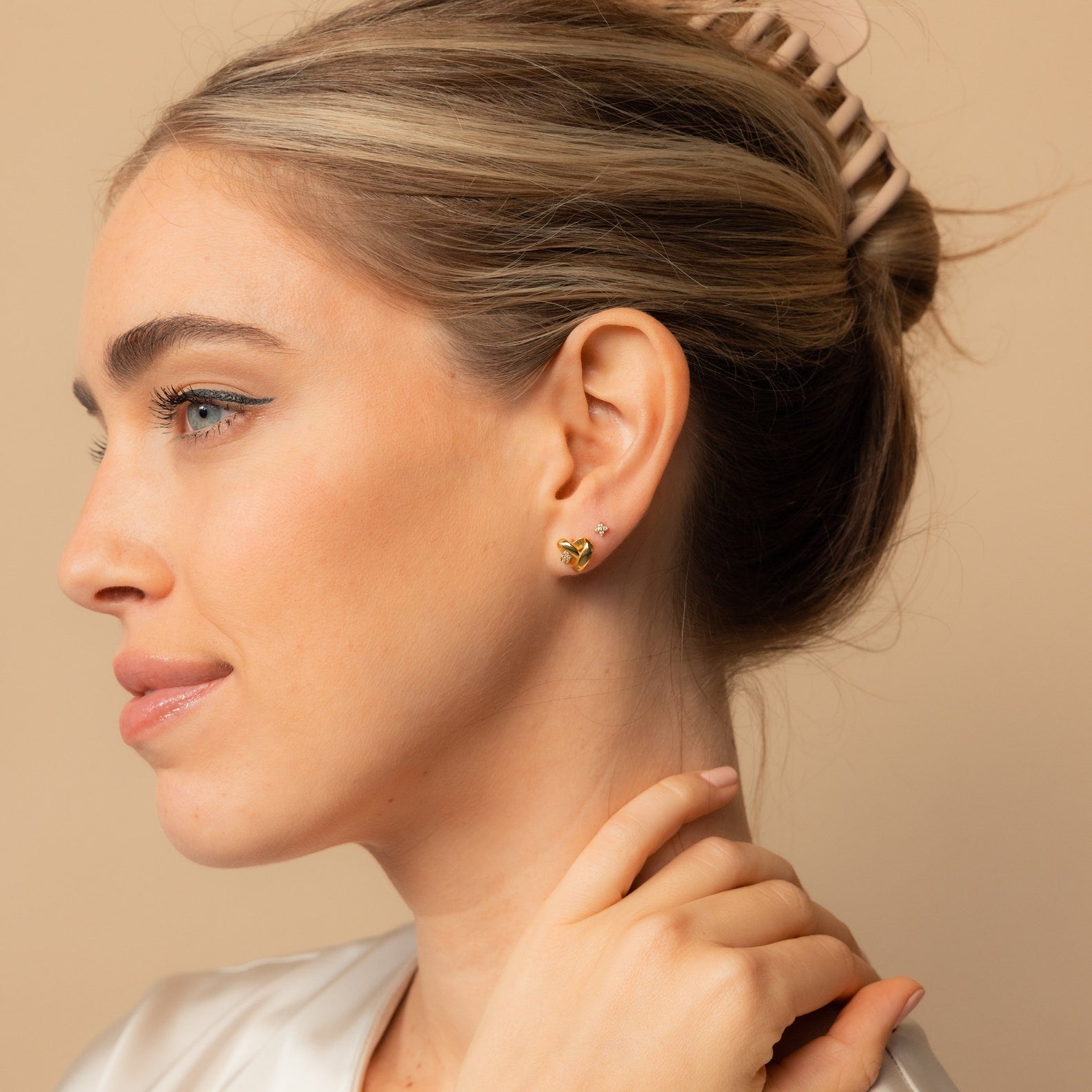A woman with light hair in a clip, wearing gold Heart Knot Studs featuring a delicate knot design, looks to the left.