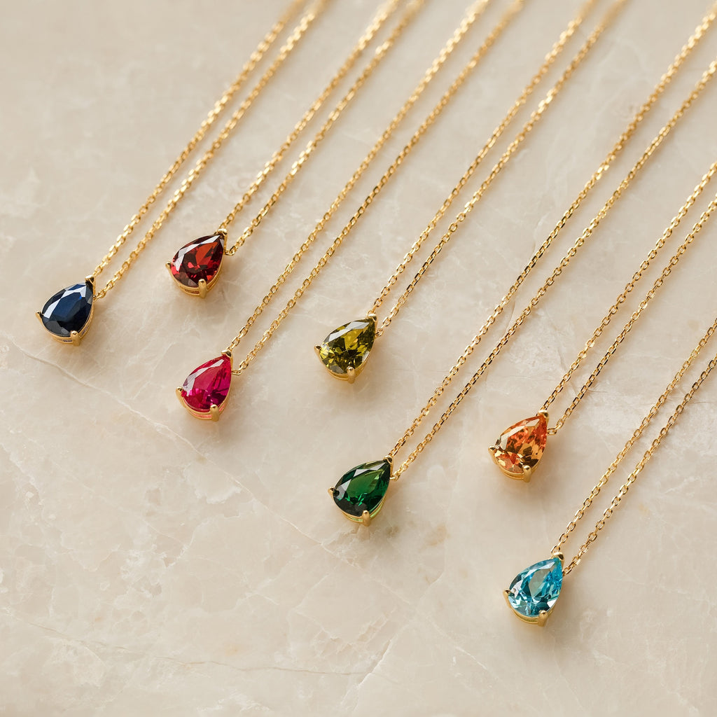 Eight Ophelia Birthstone Necklaces with gold chains and teardrop-shaped colorful gemstone pendants are displayed on marble, perfect for adding an elegant touch as a personalized birthstone accessory.