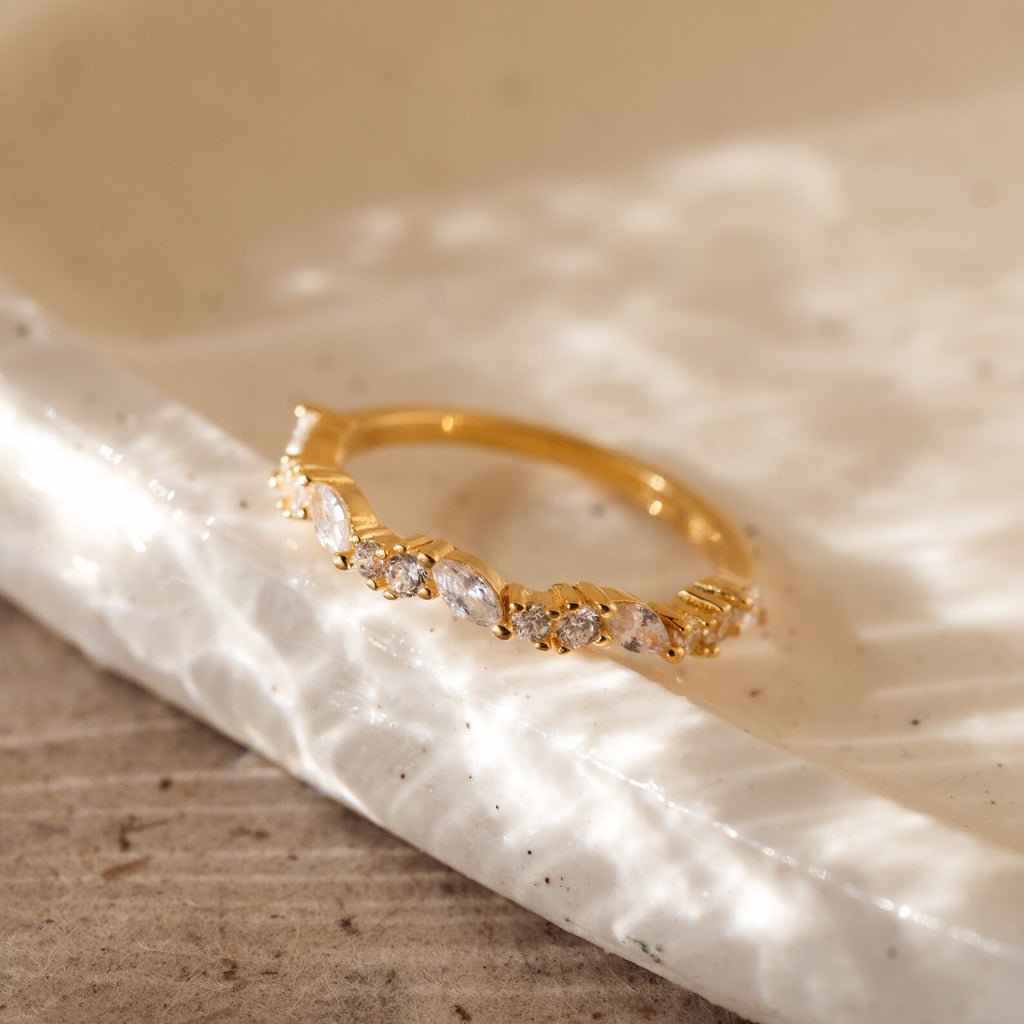 The Verity Diamond Ring, a gold cluster band adorned with sparkling diamonds, rests on a textured white ceramic dish.