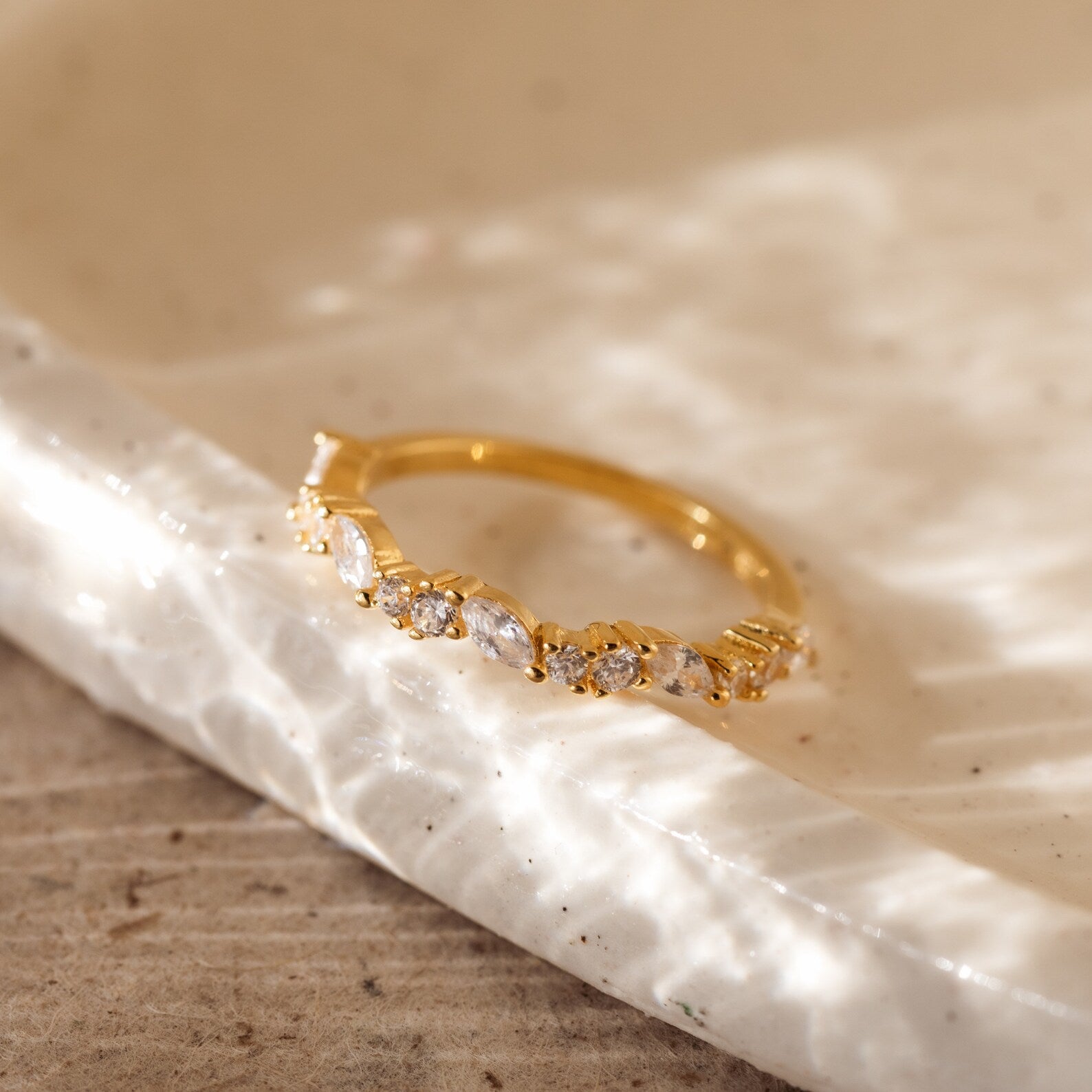 The Verity Diamond Ring, a gold cluster band adorned with sparkling diamonds, rests on a textured white ceramic dish.